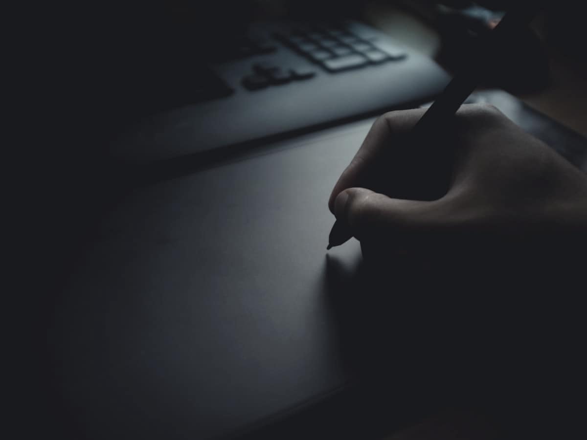 Una mano escribiendo sobre una mesa a oscuras, iluminada solo por la pantalla